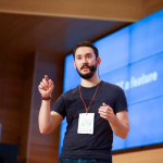 Headshot photo of Adrián Bolonio. Adrián is shown hands raised speaking at a conference