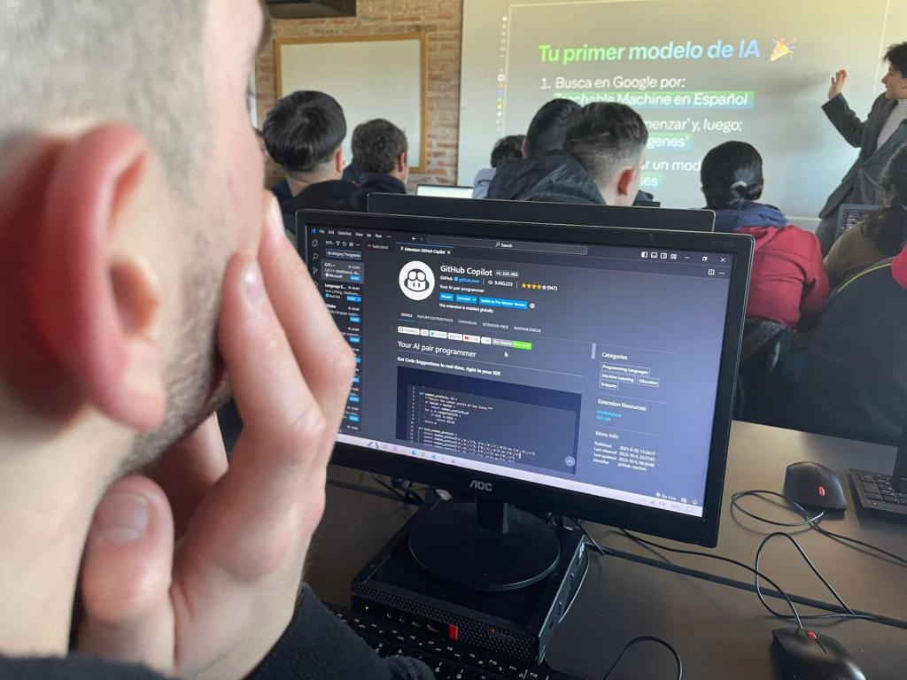 Photo of a male student taken from behind. He is resting is cheek on his right hand, and the computer screen in front of him is on a website about GitHub Copilot. Further in the distance, there is a teacher gesturing toward a projector screen displaying text in Spanish.